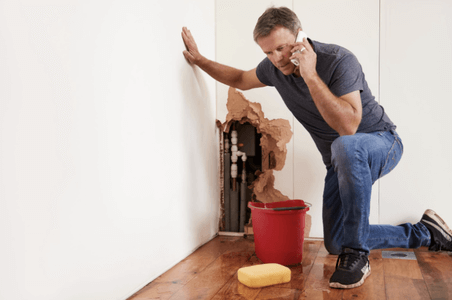 man with a bucket on the phone, water all over the floor and a ripped open wall